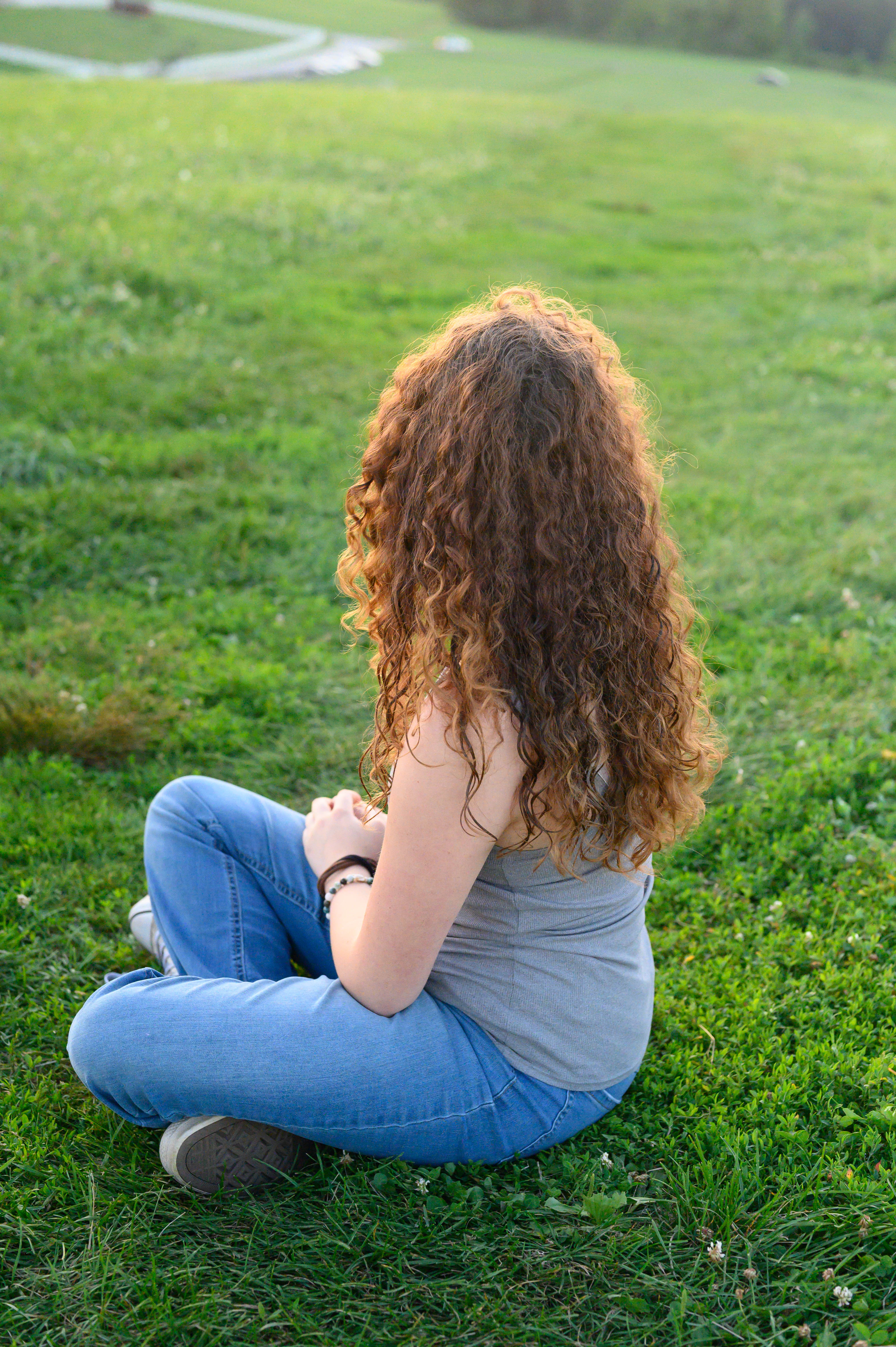 A girl with curly brown hair sitting in a field with her legs crossed while looking into the sunset